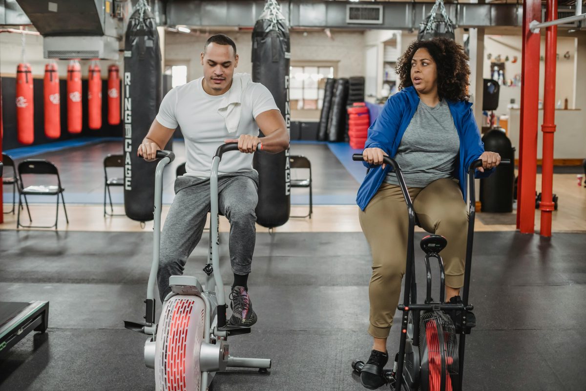 Full body of overweight black woman sitting on cycling machine and looking at fitness trainer