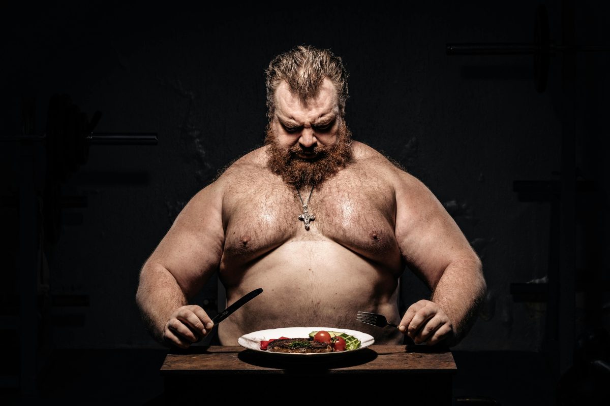 Strong Man Sitting at Table Eating Meat on Black Background